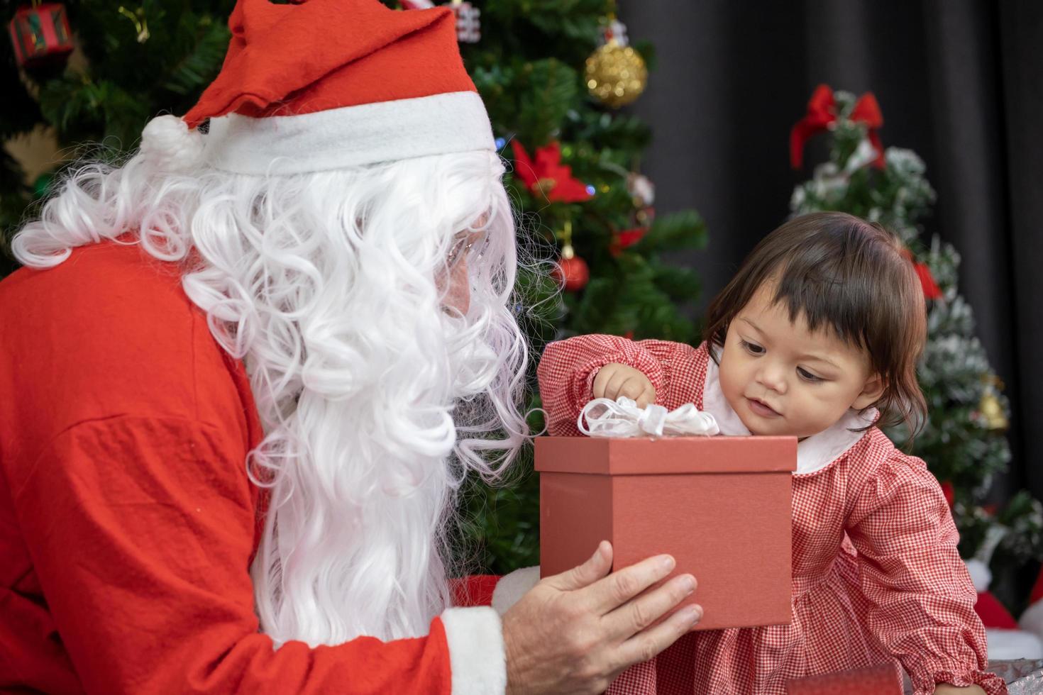 el bebé caucásico está recibiendo regalos de santa claus por la noche junto al árbol de navidad completamente decorado para el uso de la celebración de la temporada foto