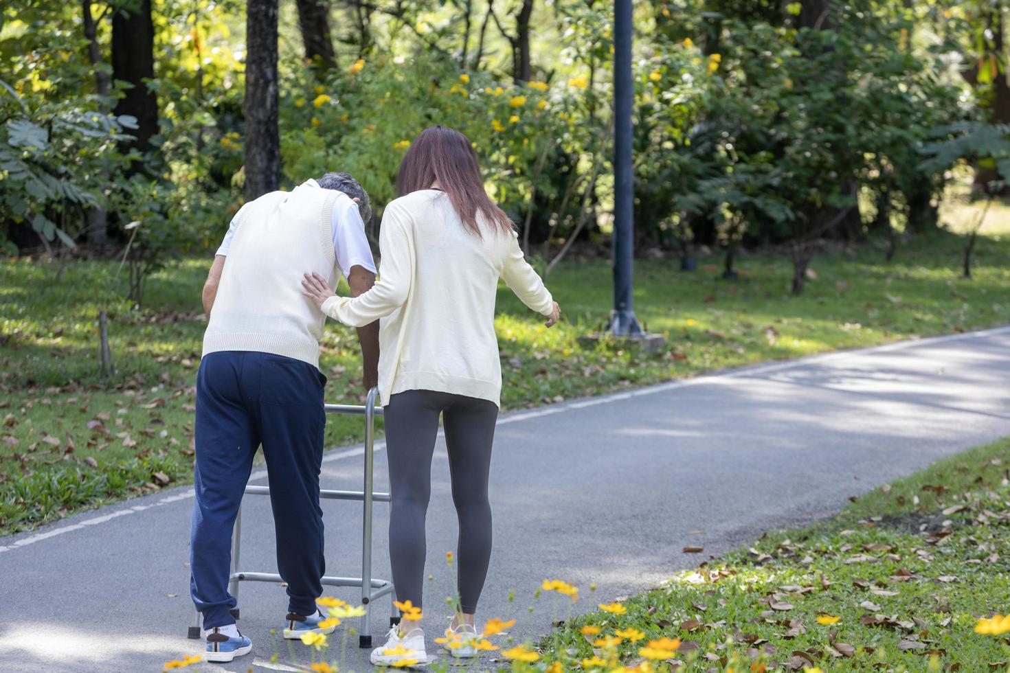 vista trasera de un padre mayor con un caminante y su hija asistente caminando juntos en el parque durante el verano para hacer ejercicio ligero y usar fisioterapia foto