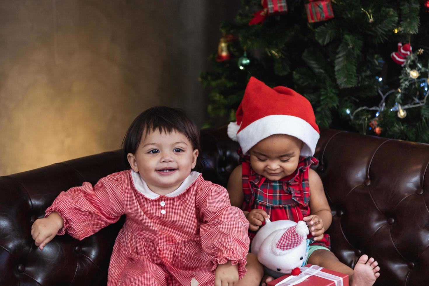 Two diverse baby friends are happily smiling while playing with present on couch dressing in christmas costume and santa hat with christmas tree on the back for season celebration concept photo