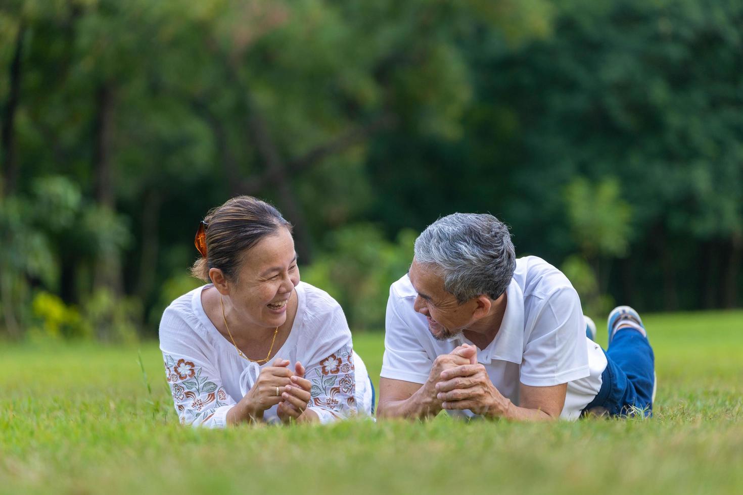 Senior Asian couple lying down on the grass in the public park laughing at each other with good mental health for longevity and happy marriage concept photo