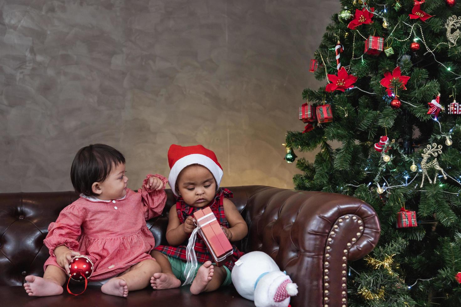 dos diversos amigos bebés sonríen felizmente mientras juegan con el presente en el sofá vistiéndose con traje de navidad y sombrero de santa con árbol de navidad en la parte posterior para el concepto de celebración de temporada foto