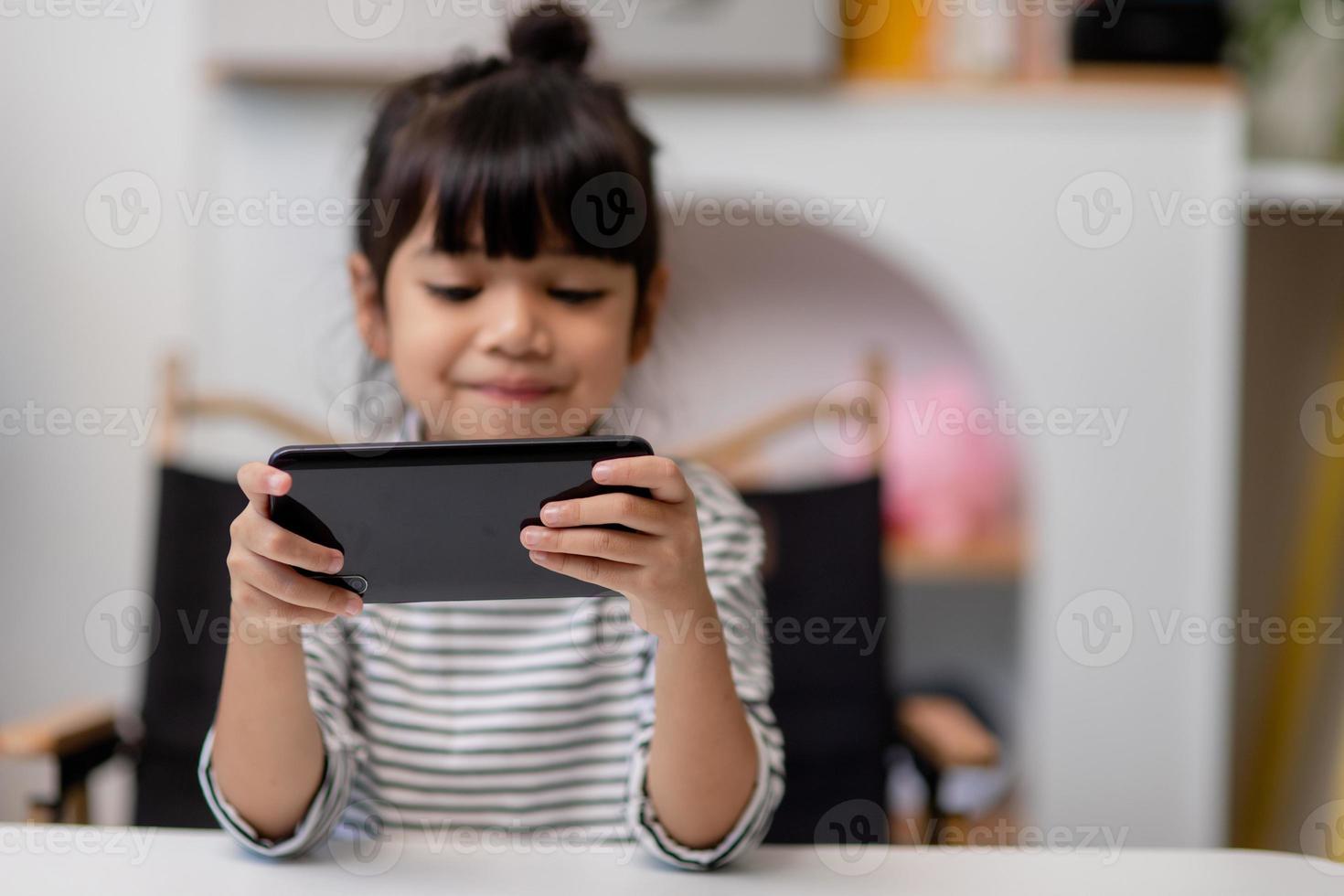 Cute little girl uses smartphone while sitting at the sofa in the living room. Child surfing the internet on mobile phone, browses through internet and watches cartoons online at home photo