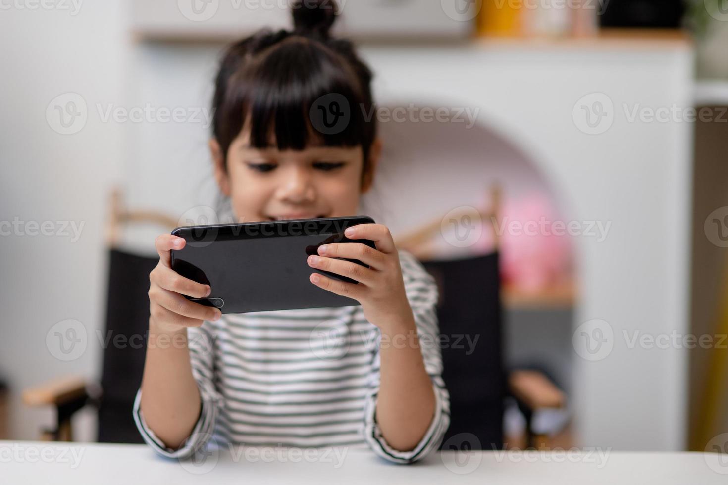 una niña linda usa un teléfono inteligente mientras se sienta en el sofá de la sala de estar. niño navegando por Internet en el teléfono móvil, navega por Internet y ve dibujos animados en línea en casa foto