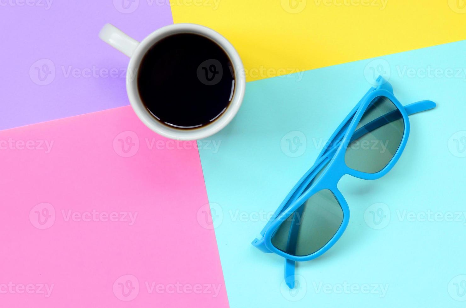 Small white coffee cup and blue sunglasses on texture background of fashion pastel blue, yellow, violet and pink colors paper in minimal concept photo