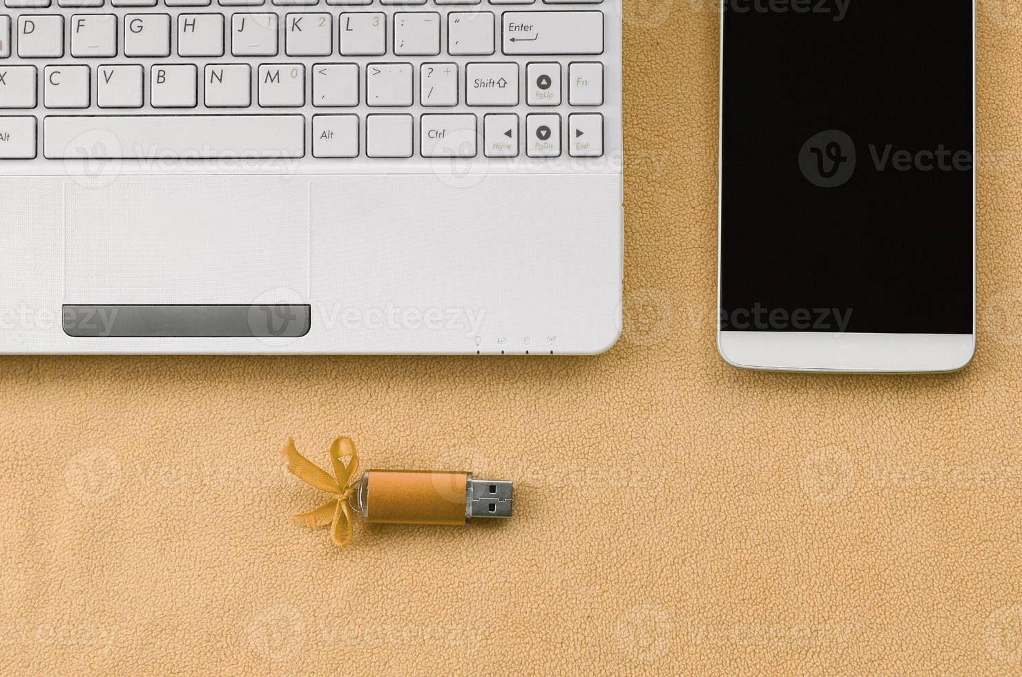 Orange usb flash memory card with a blue bow lies on a blanket of soft and furry light orange fleece fabric beside to a white laptop and smartphone. Classic female gift design for a memory card photo