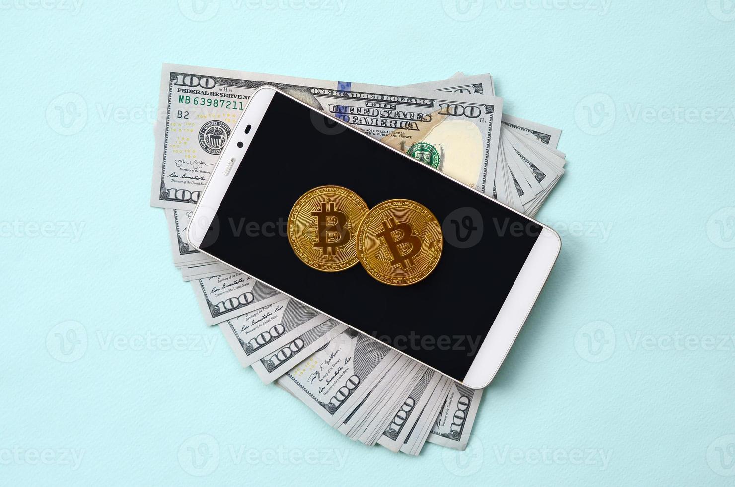 Bitcoins lies on a smartphone and hundred dollar bills on a light blue background photo