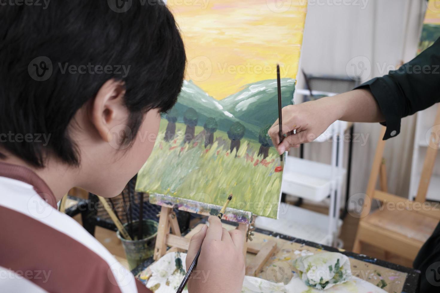 A young Asian boy concentrates on acrylic color picture painting on canvas with student kids in an art classroom, creative learning with talents and skills in the elementary school studio education. photo