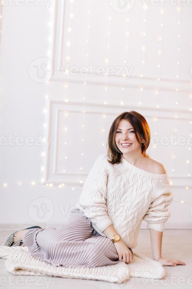 bella joven con un suéter blanco, falda y tacones altos sobre un fondo de paredes blancas y guirnaldas navideñas foto