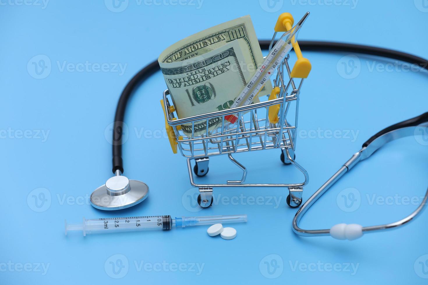 Dollars banknote and thermometer in shopping cart, white pills, syringe and stethoscope on blue background. Medicine concept with copy space. selective focus. photo