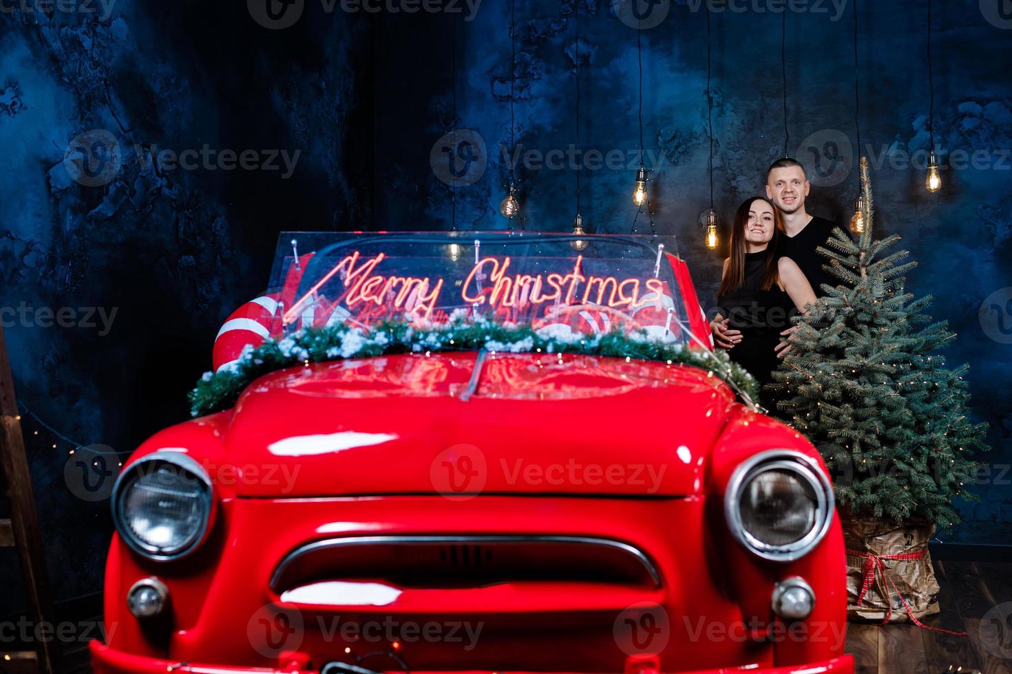 Young happy couple man and woman in love in Christmas are hugging near a red retro car with New Year's gifts and christmas tree. Kiss, girl, happiness, quarantine Christmas celebration, holiday photo