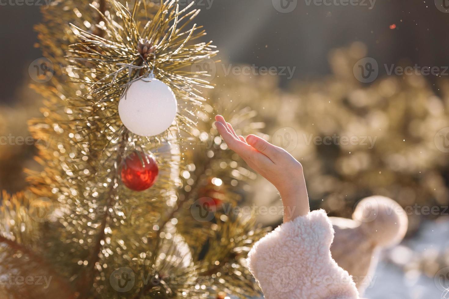 Child girl is decorating Christmas tree with shiny white and red balls outdoors before the holidays. Kid in warm jacket is having fun in park in winter day. Merry Xmas and happy family holidays photo
