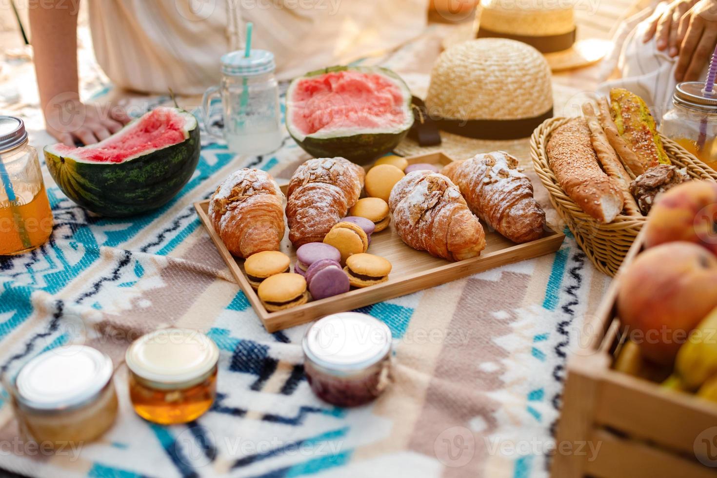 Beautiful summer picnic on the beach. Organic fresh fruit, croissants, colourful macarons or macaroons, watermelon, cold drinks and jam on boho style blanket and straw hat. photo