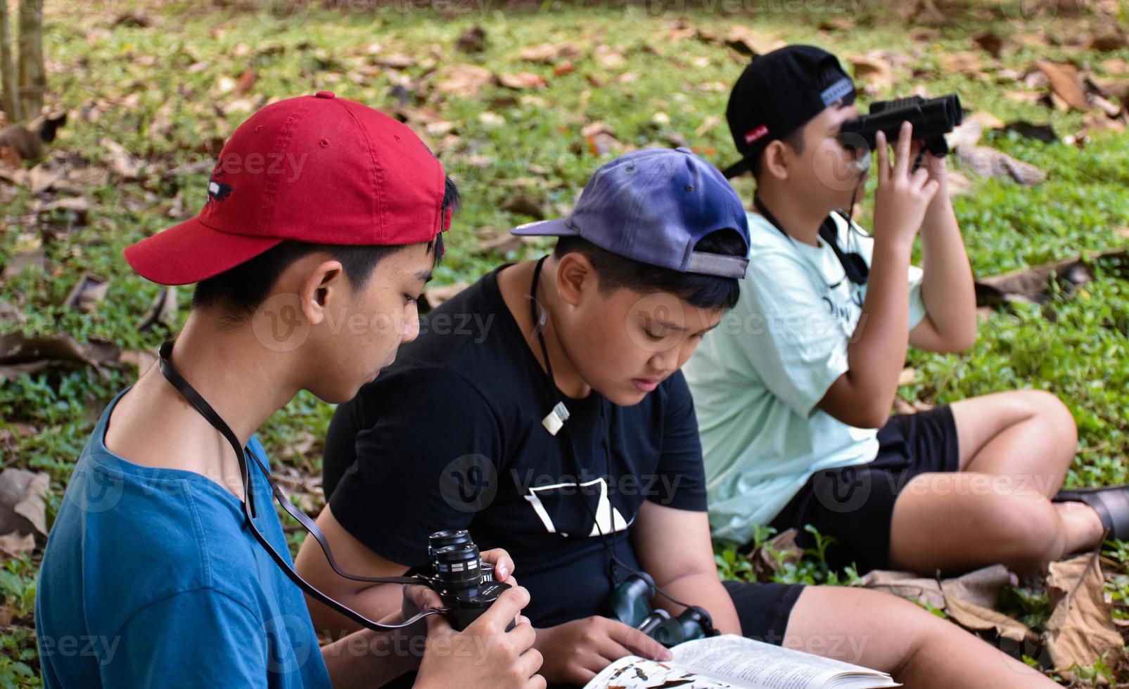 Three asian boys're reading birds details and going to use binoculars to watch birds on the trees during summer camp, idea for learning creatures and wildlife animals outside the classroom. photo