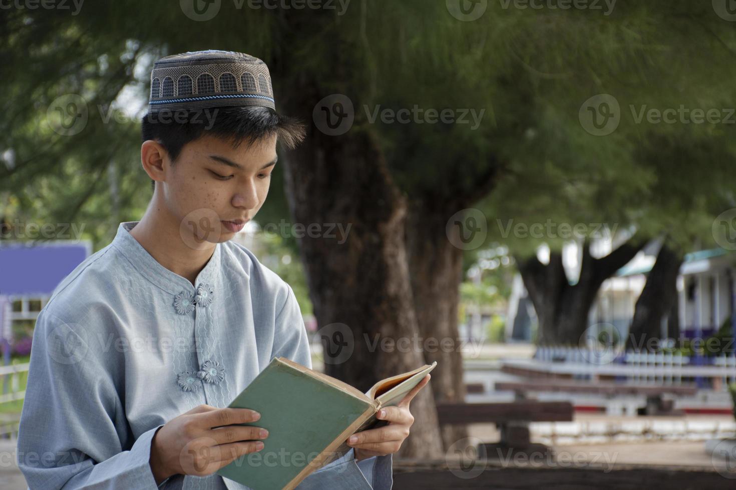 Young asian muslim boy wears hat, sitting in school park and reading his book in his free times before going back home, soft and selective focus. photo