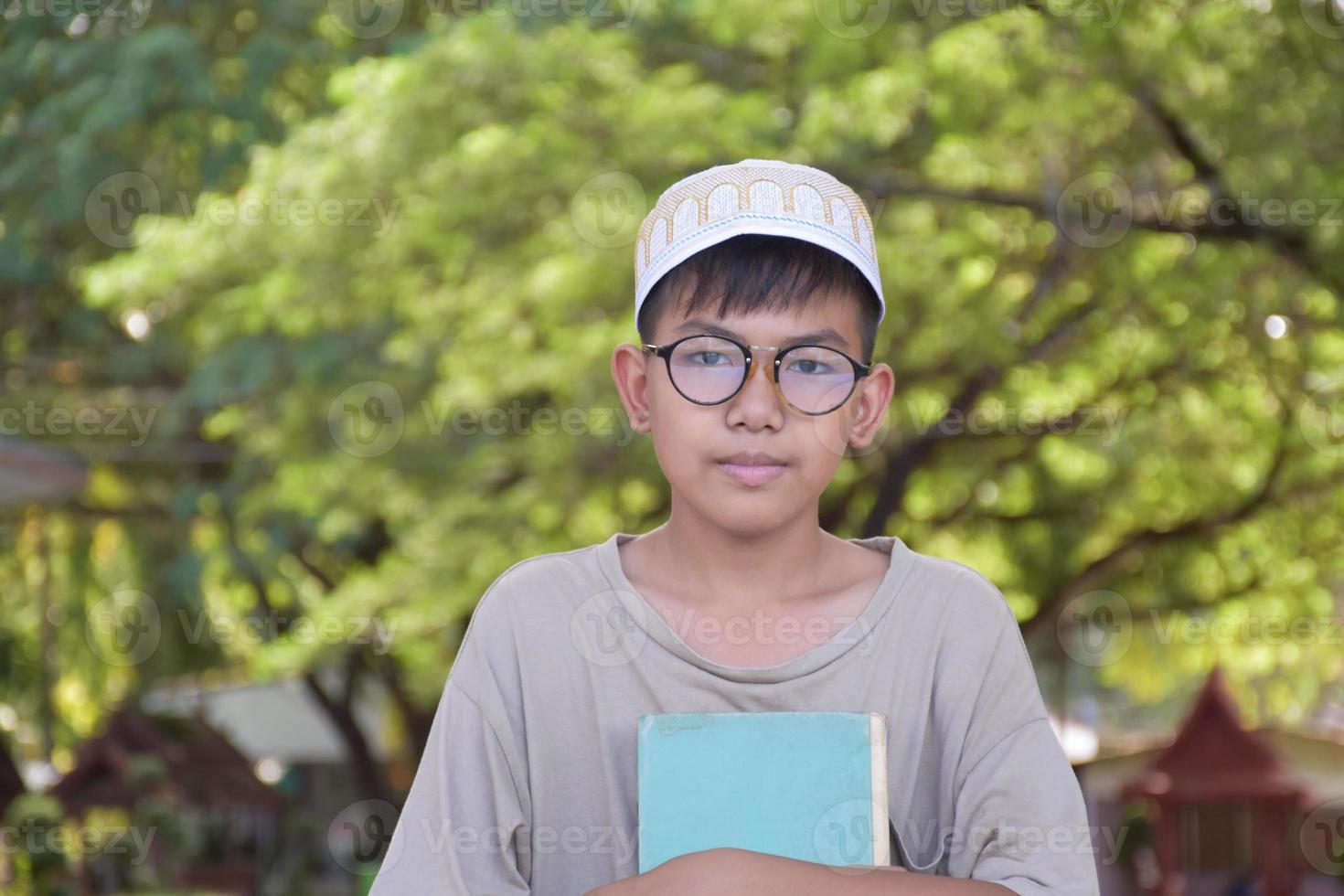 Young asian muslim boy wears eyeglasses, sitting in school park and reading his book in his free times before going back home, soft and selective focus. photo