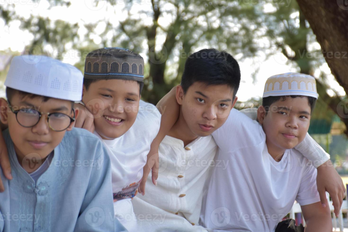 Young asian Muslim boys are playing with their hands behind each other by sitting in a row happily under a tree in the school park, soft and selective focus. photo