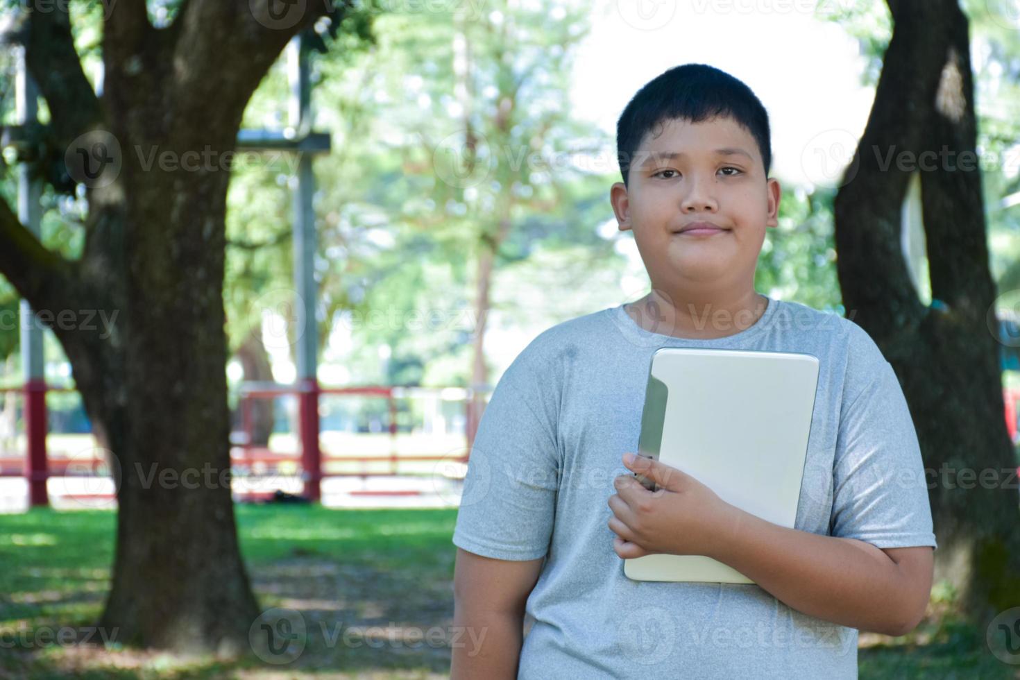 Portrait young asian boy holds laplet and stands under the big tree, soft focus, concept for using technology in daily life. photo