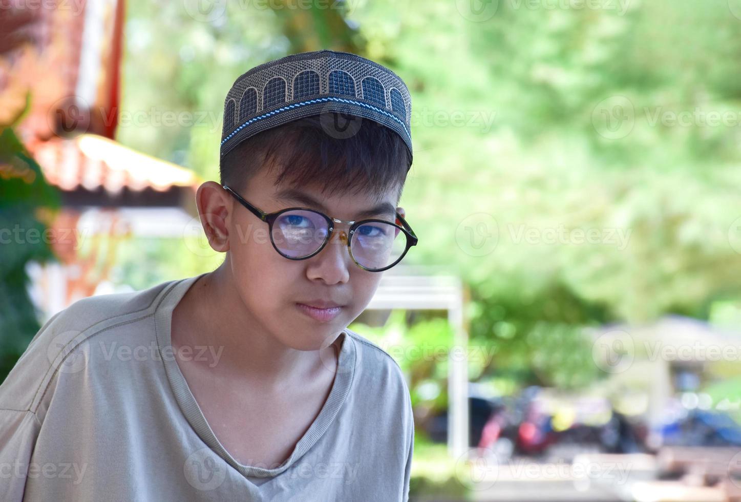 Young asian muslim boy wears eyeglasses, sitting in school park and reading his book in his free times before going back home, soft and selective focus. photo