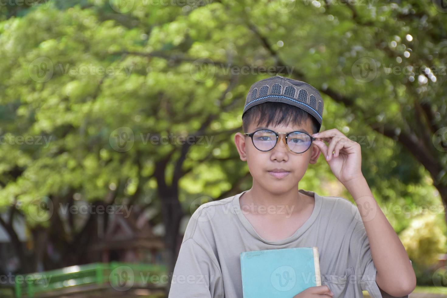 Young asian muslim boy wears eyeglasses, sitting in school park and reading his book in his free times before going back home, soft and selective focus. photo