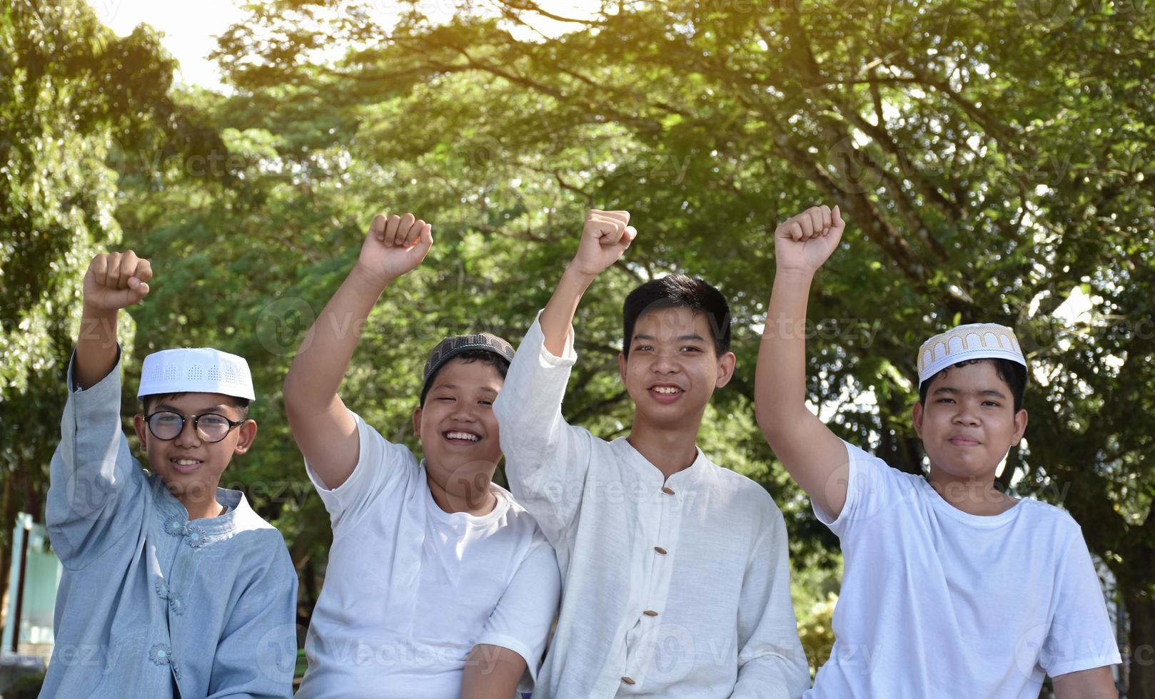Young asian Muslim boys are raising their right hands to show their victory and success happily under a tree in the school park, soft and selective focus. photo