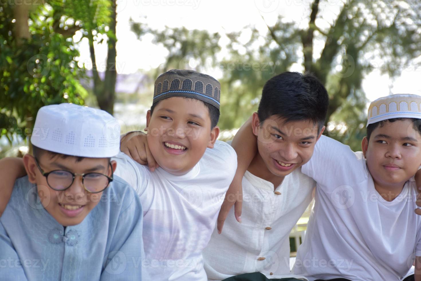 Young asian Muslim boys are playing with their hands behind each other by sitting in a row happily under a tree in the school park, soft and selective focus. photo