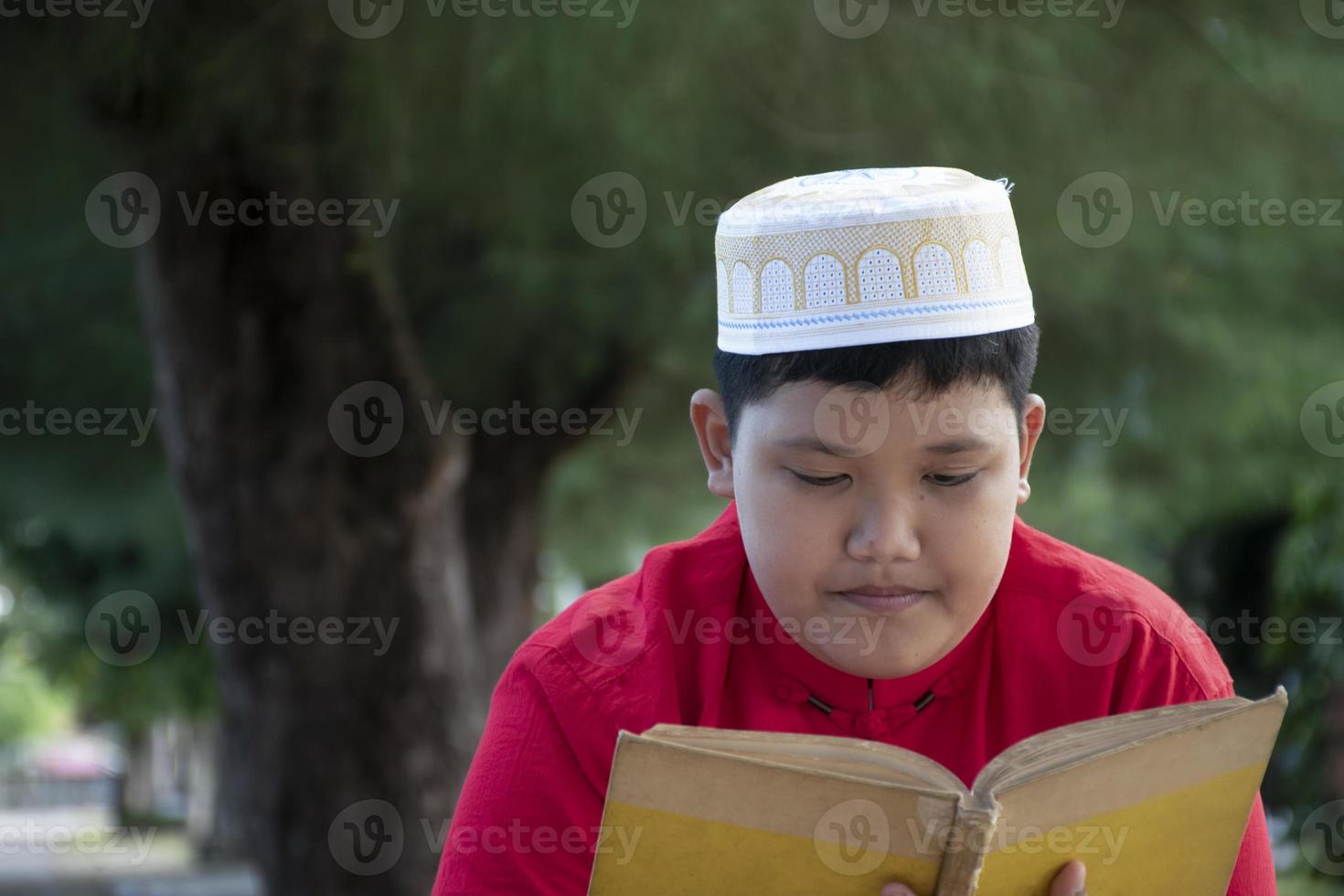 Young asian muslim boy wears hat, sitting in school park and reading his book in his free times before going back home, soft and selective focus. photo