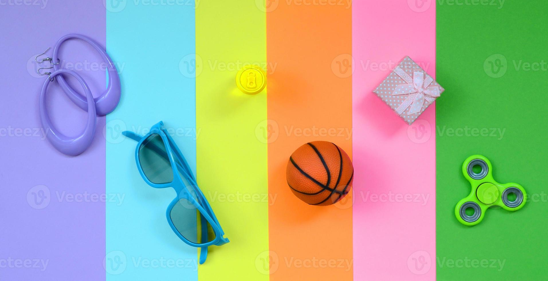 composición pastel de moda con aretes, gafas de sol, lata de bebida, pelota de baloncesto, caja de regalo y spinner sobre fondo de colores rosa, violeta, verde, naranja, amarillo y azul foto