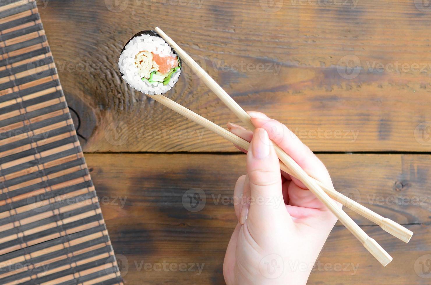 A hand with chopsticks holds a sushi roll on a bamboo straw serwing mat background. Traditional Asian food photo