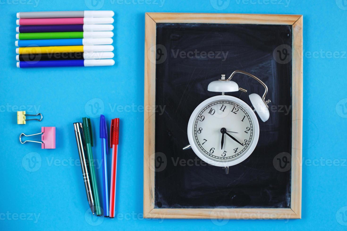 pizarra escolar y accesorios de papelería, lápices, reloj despertador y bolígrafos sobre fondo azul. concepto de regreso a la escuela. foto