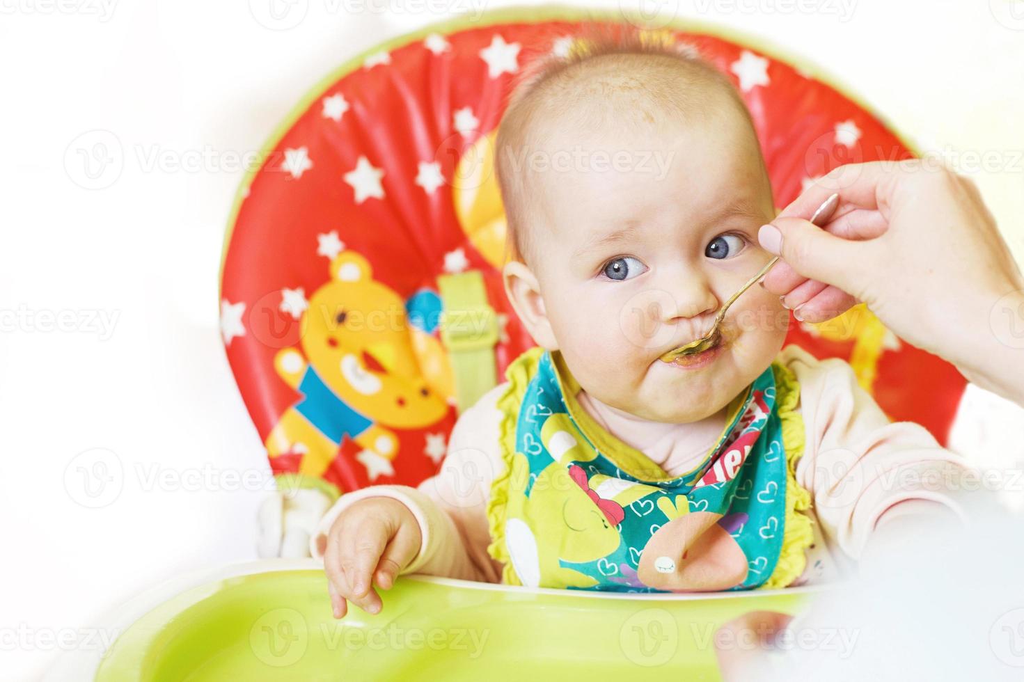 Mom feeds funny baby from a spoon on a white background. child eats in a highchair photo