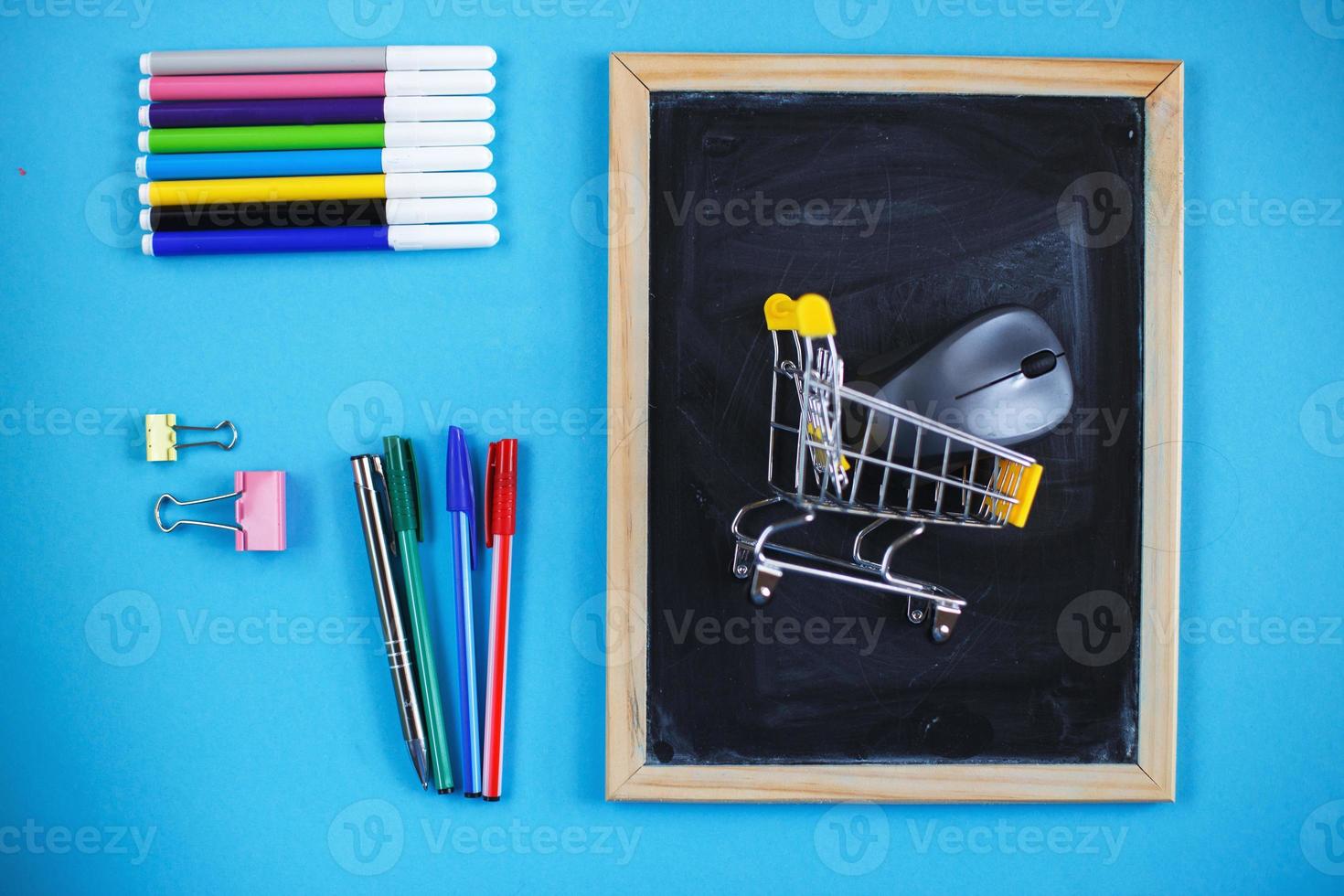 School chalkboard and stationery accessories, pencils, pens, shopping cart on blue background. Back to school concept. photo