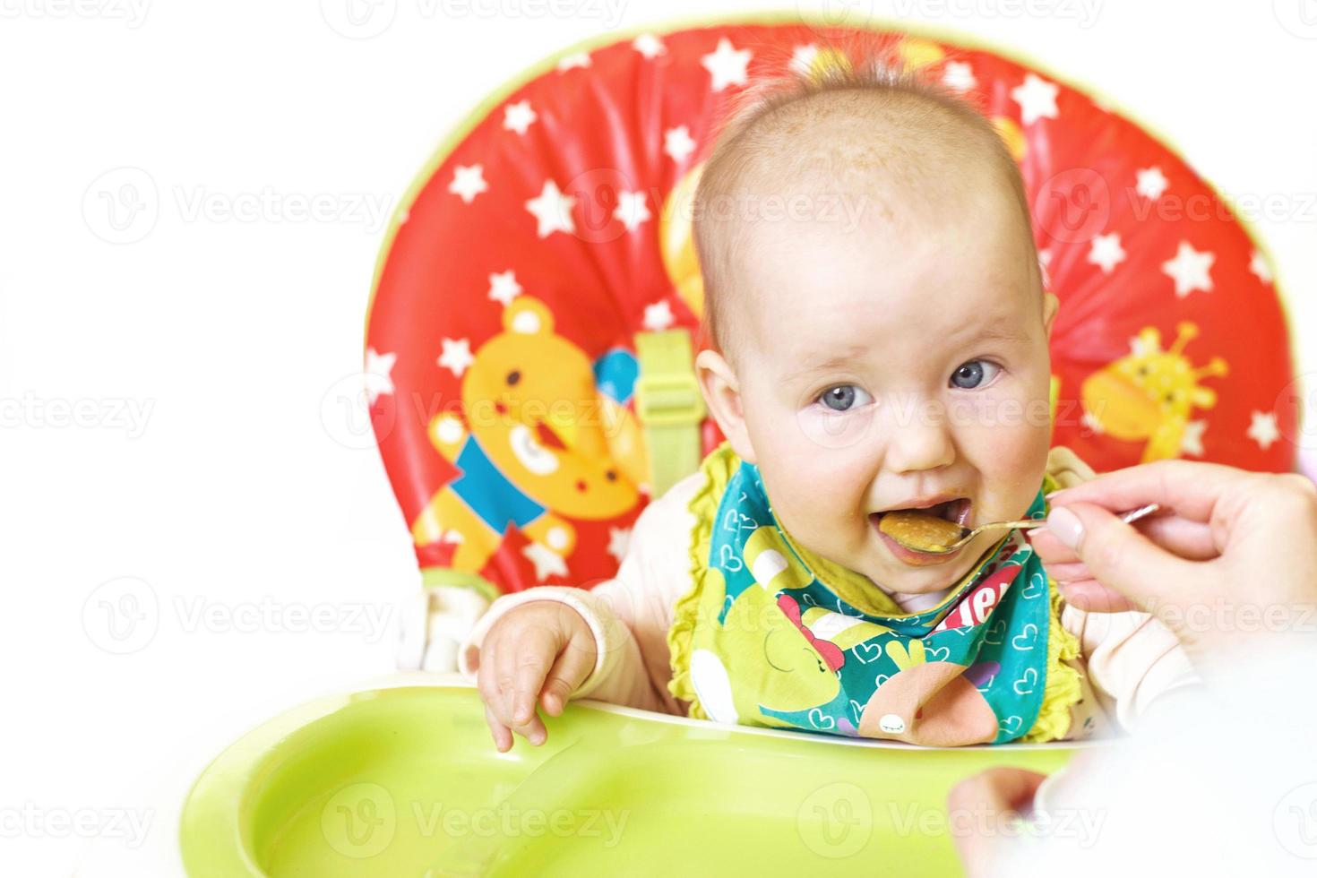 Mom feeds funny baby from a spoon on a white background. child eats in a highchair photo