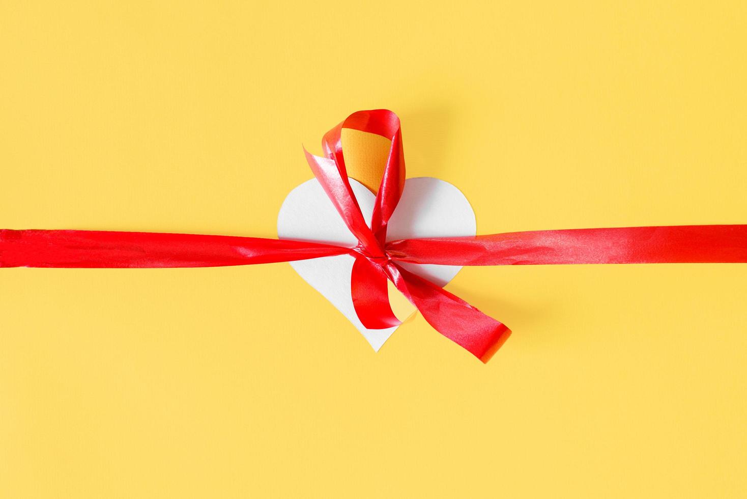 White paper heart and red bow on a yellow background, top view. Creative idea to give love photo