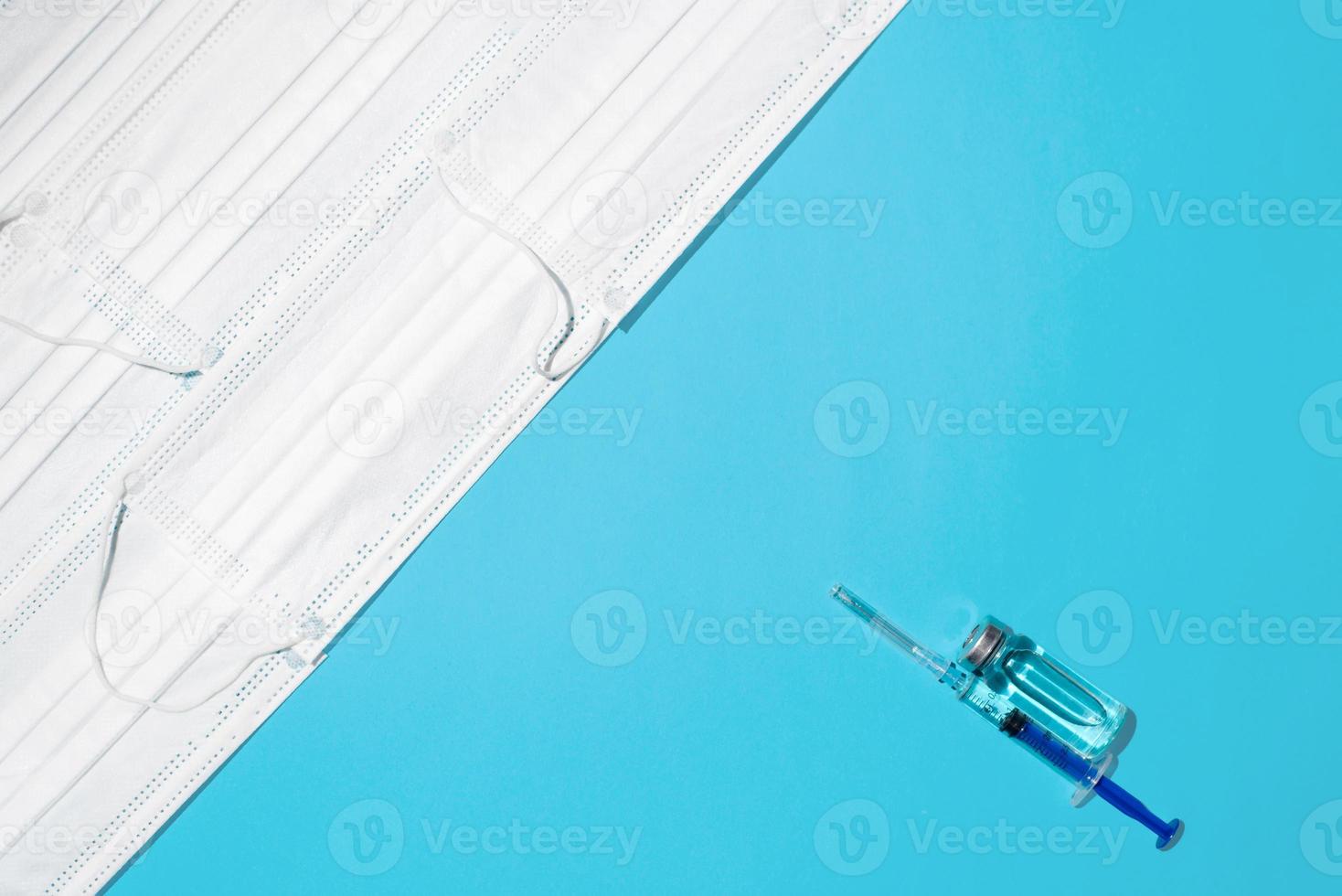 Concept war against virus, vaccination of population. Vaccine vial, syringe and bunch of medical face masks on blue background, top view photo