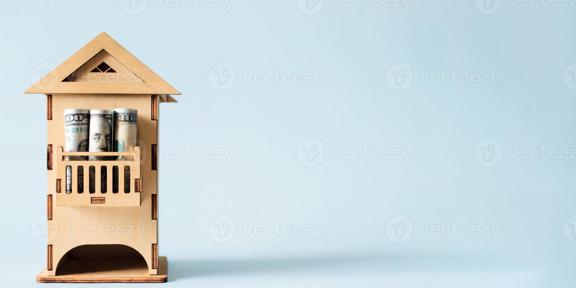 Wooden toy house and one hundred dollar bills on a light blue background. Concept for real estate prices, apartment rental, mortgage, construction business. Copy space banner photo