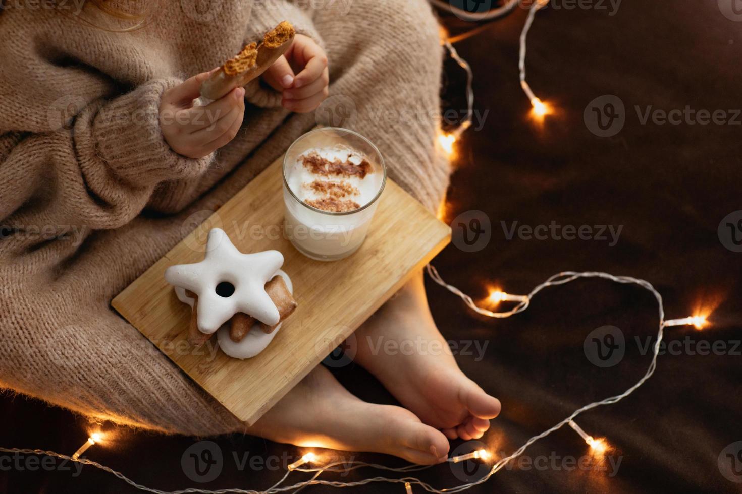 los pies y las manos descalzos del niño sostienen un vaso de galleta de jengibre con ponche de leche, dulces navideños galletas de jengibre. luces bokeh. foto