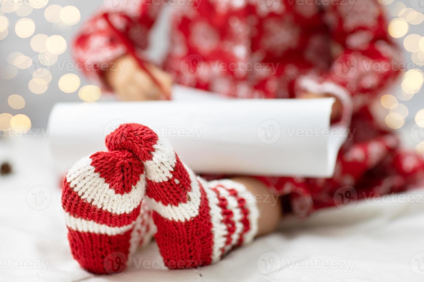 los pies de los niños se acercan y los calcetines rojos y blancos de punto de lana. niña escribiendo una carta de navidad lista de deseos para santa claus en una habitación decorada festivamente. foto