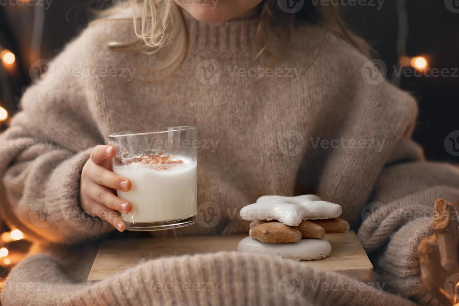 los pies y las manos descalzos del niño sostienen un vaso de galleta de jengibre con ponche de leche, dulces navideños galletas de jengibre. luces bokeh. foto