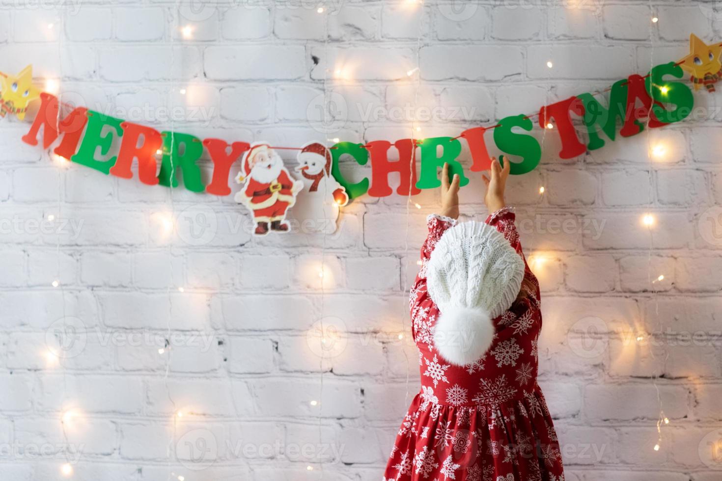 Child girl in knitted hat with pompon decorated break white wall at home garland Merry Christmas and lights photo