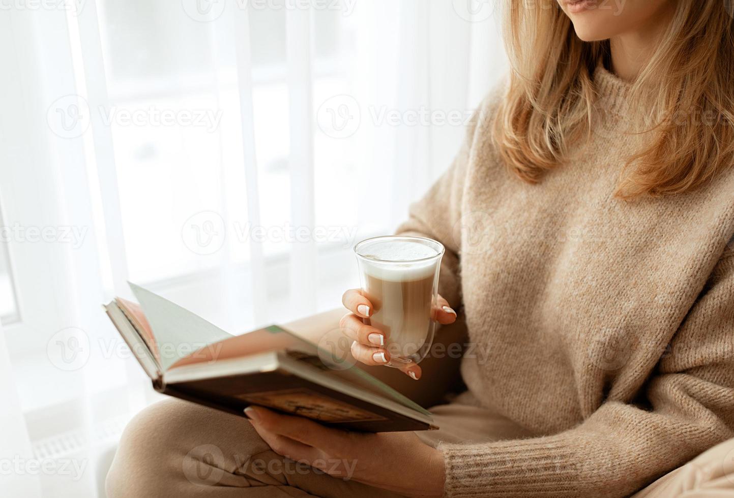 Close up women's hands and fingers with a beautiful French manicure holding cup transparent glass with coffee with white foam.Copy space for text, banner. Beige knitted sweater and background. photo