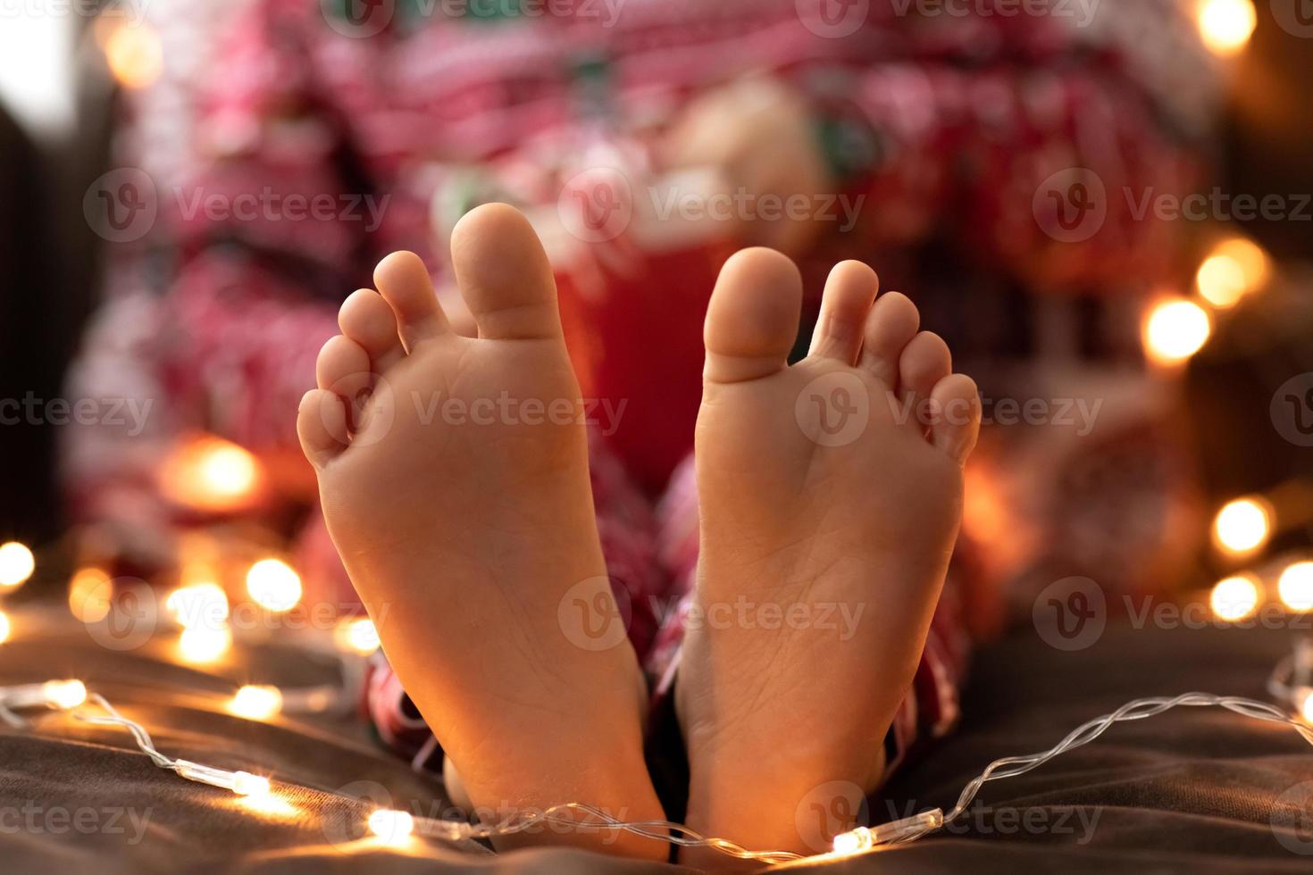 Close up Child bare feet Kid in red Christmas pajamas hold mug cup striped green red candy Christmas canes bokeh lights background. New year photo