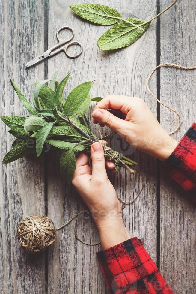 Sage leaf bundle in hands, hemp string and scissors on old wooden table photo
