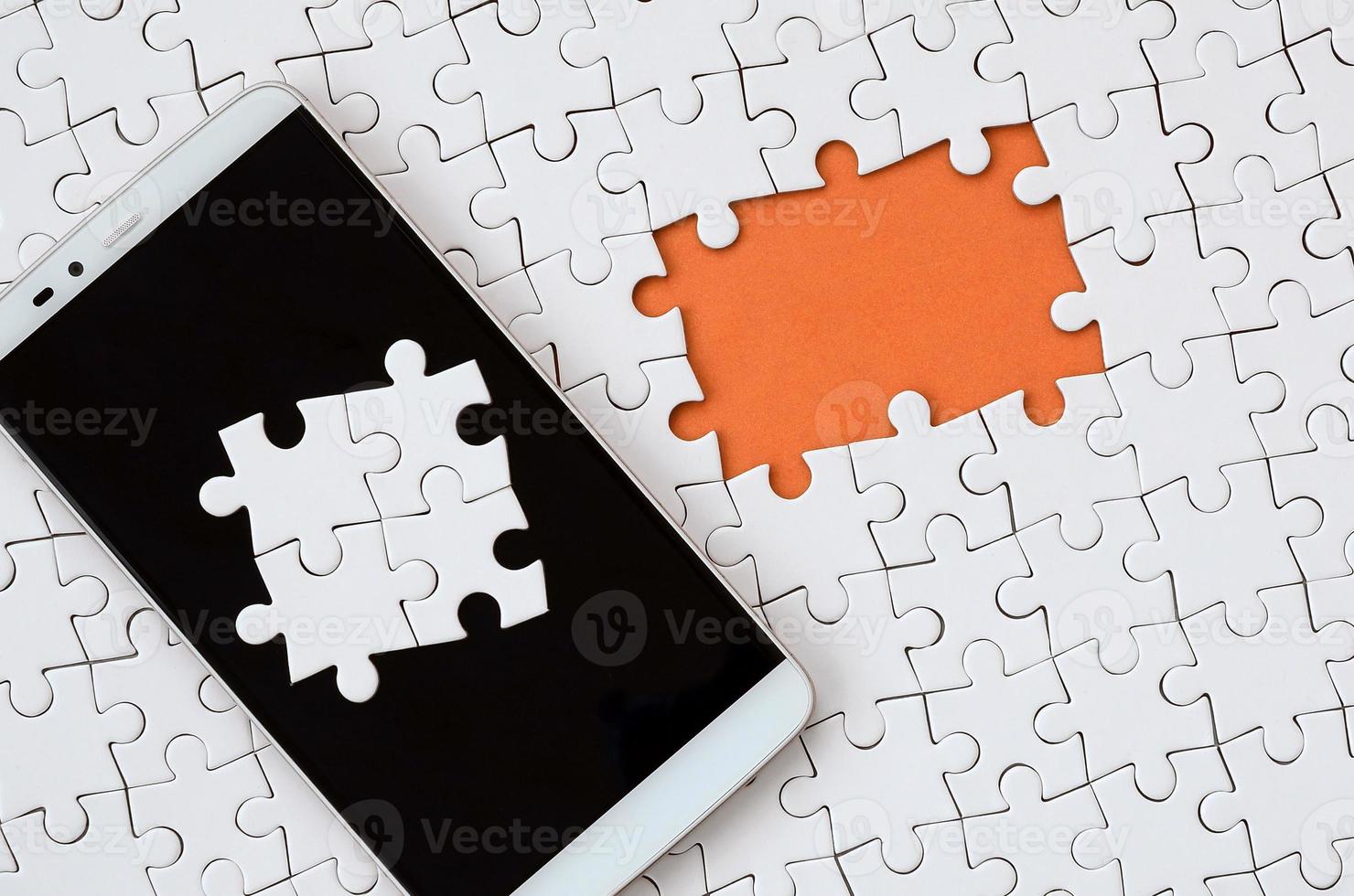 A modern big smartphone with several puzzle elements on the touch screen lies on a white jigsaw puzzle in an assembled state with missing elements photo