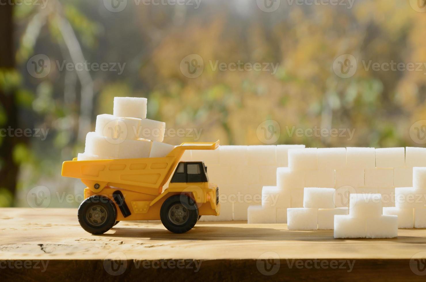 A small yellow toy truck is loaded with white sugar cubes near the sugar wall. A car on a wooden surface against a background of autumn forest. Extraction and transportation of sugar photo