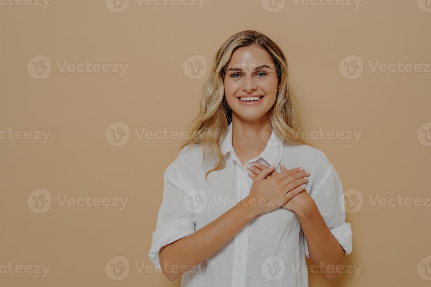 Young woman in love holding hands on chest, gazing at camera as feeling grateful and appreciative photo