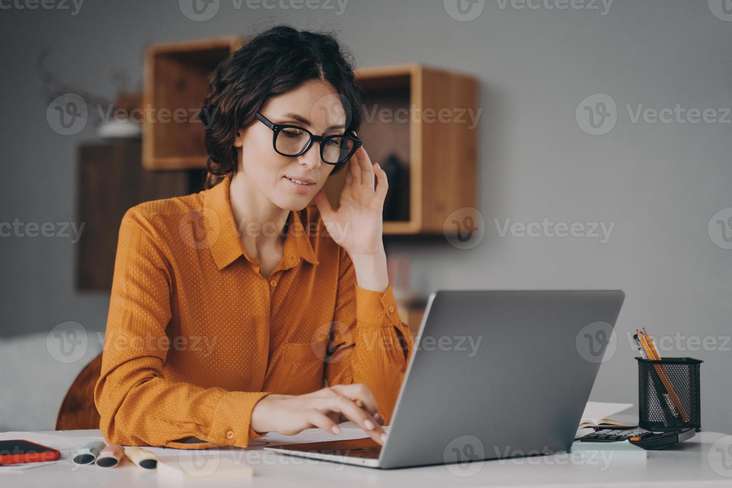 mujer española enfocada con anteojos se sienta en el escritorio y escribe en la computadora portátil durante el trabajo remoto en casa foto