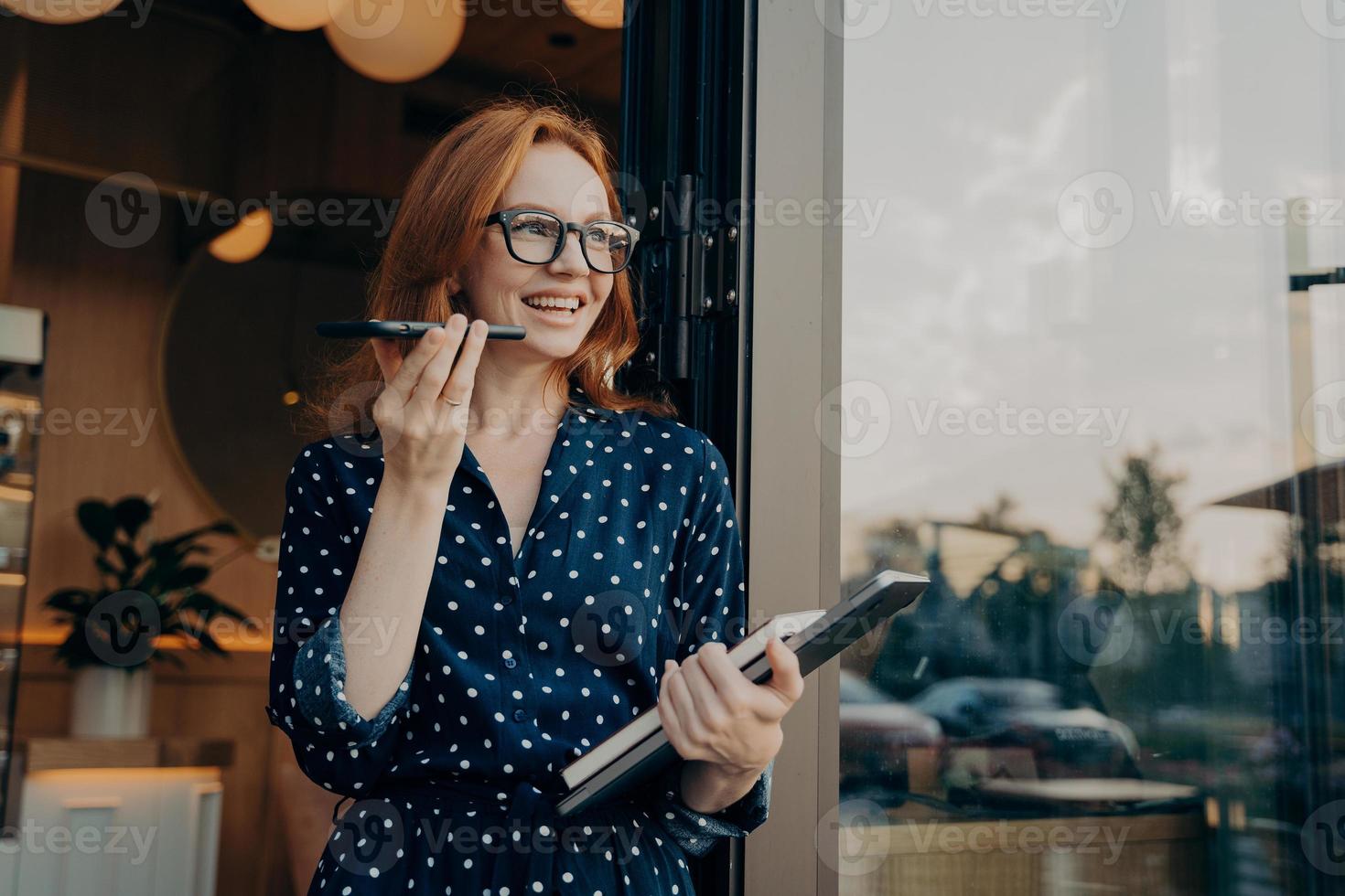 Happy ginger business woman freelancer with laptop holds mobile phone and talks on speakerphone photo