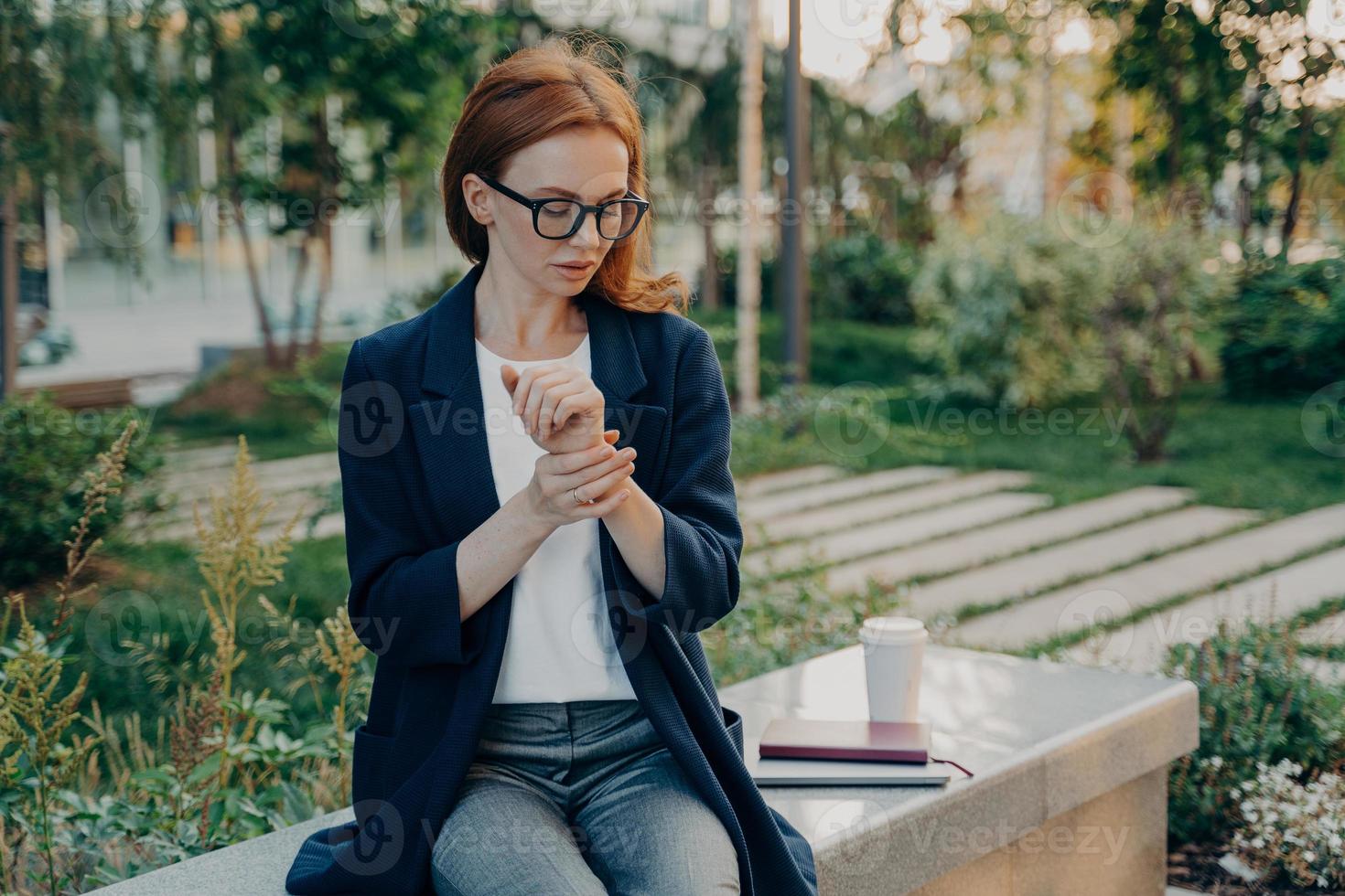 Young tired redhead business woman measuring pulse on wrist, checking heart rate, sitting in park photo