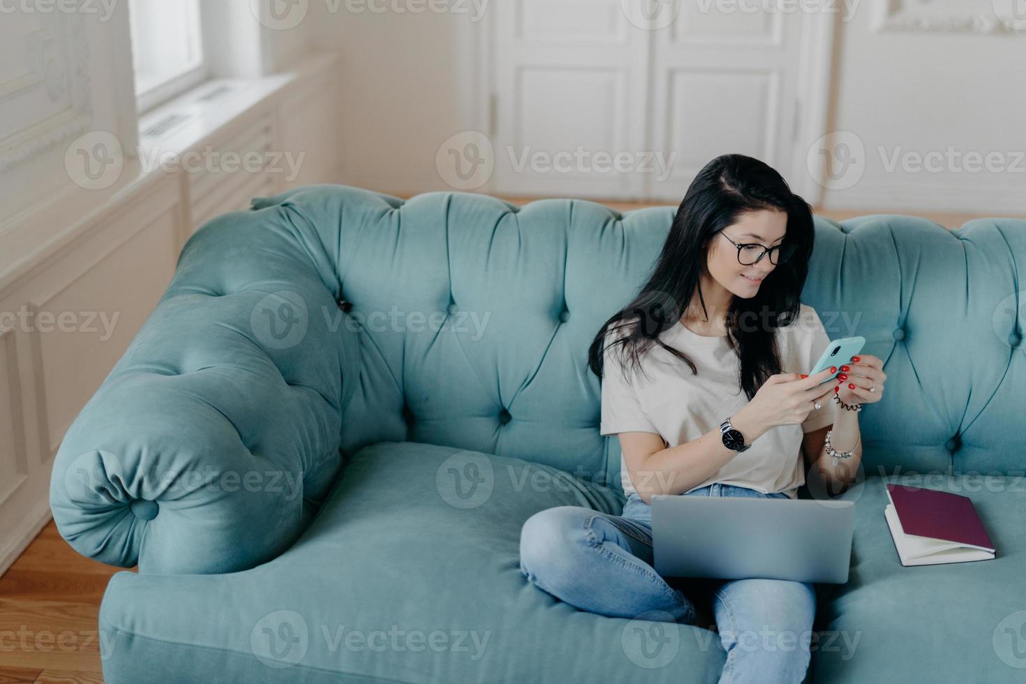 View from above of busy female freelancer sits in cozy room on comfortable sofa, works on new startup project, uses laptop computer, holds mobile phoneand sends text messages, interacts with netbook photo