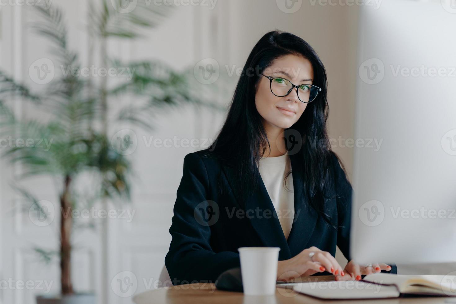 Serious successful female office worker in formal outfit, wears eyeglasses, busy with online work, poses at desktop, works on computer, reads business new or watches tutorial, enjoys working process. photo